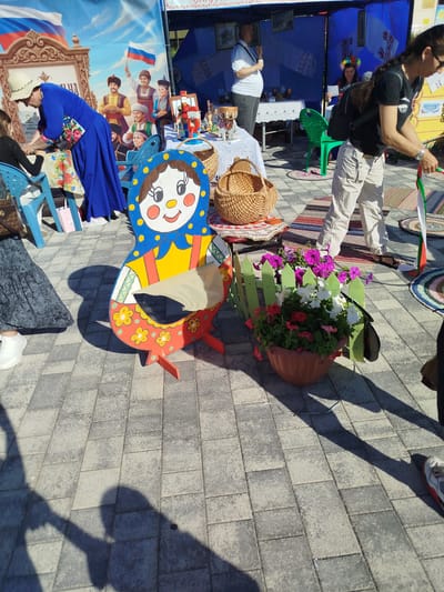 The image captures an outdoor scene at what appears to be a cultural event or market, possibly in Zavodoukovsk, Russia. In the foreground, a large, colorful, matryoshka doll-shaped chair is prominently displayed. It is painted with a cheerful, stylized face, a yellow shirt, and a red apron adorned with floral patterns. The chair is situated on a paved area with a shadow of the doll falling across it. To the right of the chair, a planter filled with vibrant pink, white, and red petunias is enclosed by a small, green wooden picket fence.
In the background, several people are visible, interacting with stalls or displays. A woman in a bright blue dress and a white hat is bending over, seemingly engaged in an activity at a table. Another person in a black t-shirt and light-colored pants is standing to the right, holding colorful ribbons. Further back, a large banner depicts several men in traditional attire and Russian flags, suggesting a patriotic or cultural theme. The setting appears to be daytime, with bright sunlight casting distinct shadows, indicating fair weather. Other objects like woven baskets, tables with items, and chairs are also present, contributing to the market-like atmosphere. The overall mood seems festive and community-oriented.