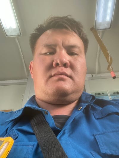 This is a selfie taken by a person wearing a blue uniform with a black strap across their chest. The person is looking directly at the camera with a neutral expression. They have short, dark hair and visible stubble on their face. In the background, there are fluorescent lights on the ceiling, and some signage with Russian text is visible on the wall. One of the signs appears to be an instructional diagram. A yellow object, possibly a piece of equipment, is partially visible in the lower right corner. The lighting suggests an indoor setting, possibly during daytime given the brightness of the lights. The presence of a uniform and the Russian text on the signage might suggest a work environment, potentially related to transportation or industrial work, given the context of "Қосшағыл ауылдық округі, Kazakhstan".
