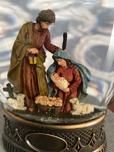 The image shows a Nativity scene figurine enclosed in a snow globe. The figurine depicts Mary holding the baby Jesus, with Joseph standing beside them holding a lantern. Two lambs are positioned at the front of the scene. The base of the snow globe is ornate and metallic. The background within the snow globe is blurry and appears to be illuminated with light and shadow. The exterior of the snow globe is transparent glass, revealing the figurine and its base. The image does not provide any information about the city of Oryol, Russia.
