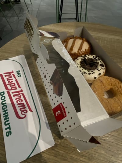 The image displays a Krispy Kreme doughnut box and a paper bag resting on a wooden table in an indoor setting, identified as Sandton, South Africa. The white Krispy Kreme "3 Doughnuts" cardboard box is partially open, revealing three distinct doughnuts inside. The top-left doughnut features light beige icing with dark stripes and a light brown crumb topping. Below it, a white-iced doughnut is decorated with dark chocolate shavings and crumbs. To the bottom-right, a plain, light brown, ring-shaped doughnut with a textured surface is visible. A white Krispy Kreme paper bag, bearing the "EAT Krispy Kreme DOUGHNUTS" logo in red and green, lies to the left of the box. The wooden table exhibits a visible grain pattern. In the blurred background, parts of green metal chair or stool legs are discernible against a light grey floor. No individuals are present.