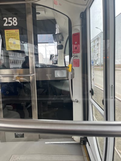 The image displays the interior of a public transport vehicle, specifically identified by a "KONCAR Electric Vehicles Inc. 2500 LT 2021" label, looking forward towards the driver's compartment and the exterior. The vehicle number "258" is visible on a transparent partition separating the passenger area from the driver's station. Affixed to this partition are a yellow sign with a QR code and Latvian text indicating "Liepāja sabiedriskajā transportā" (Liepaja public transport) and "BIĻETES VAI REĢISTRĀCIJA" (TICKETS OR REGISTRATION). A silver vertical grab handle with a yellow activation button and a red emergency panel is located on the right side of the partition. Through the partition, multiple electronic screens are visible within the driver's compartment, and a dark reflection suggests the presence of a driver. The exterior scene, visible through the vehicle's windows, shows an urban street in Liepaja, Latvia, under an overcast sky. Features include a grey, multi-story residential or institutional building, a street light pole, and a blue informational sign with a wheelchair symbol on the right-side window.
