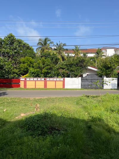 The image depicts a sunny day in Kalutara, Sri Lanka, with a clear blue sky dotted with wispy clouds. A road runs horizontally across the lower half of the frame, bordered by a vibrant, multi-colored wall. The wall features sections of bright red, yellow with decorative white patterns, and plain white. Behind the wall, lush green trees and palm trees dominate the landscape, interspersed with glimpses of residential buildings. One building, visible behind the central section of the wall, has a red tiled roof and solar panels. Further to the right, a modern white building with solar panels is also visible. A dark metal gate, intricately designed, stands out against a white wall to the right of the yellow section of the boundary wall. The foreground is covered in thick, green grass, with a narrow path of trampled grass suggesting a natural walkway. The overall scene conveys a peaceful, tropical residential environment.