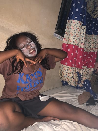 A young Black woman is lying on the floor with a white facial mask on. She wears a brown t-shirt with blue lettering and black shorts. She is making a peace sign with her hand. A bed is visible behind her, covered in a patterned comforter. The floor is beige. There is no information to identify the location in Agban, Nigeria.