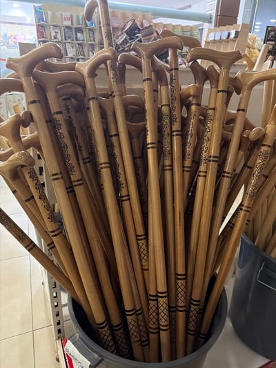 The image displays a large collection of wooden walking canes with decorative patterns, clustered together in a grey bucket. The canes are predominantly light brown and have curved handles. Some of the canes feature black patterned designs, including floral motifs and geometric shapes, around sections of their shafts. The background suggests an indoor retail environment, possibly a shop, with shelves displaying books and other items in the upper left corner. The floor appears to be tiled. The specific location is in Acıpayam, Turkey.