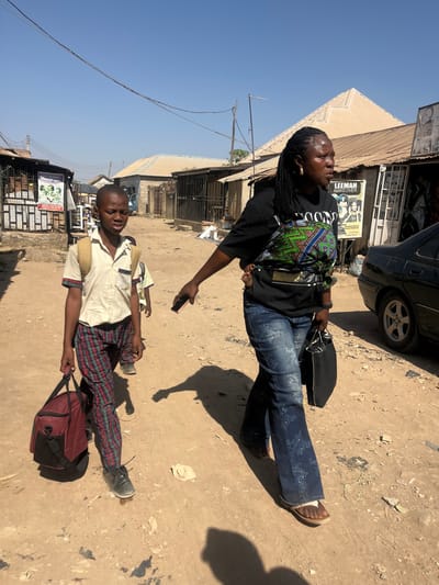 The image depicts a daytime scene on an unpaved, dusty dirt road in the city of Dzhidu, Nigeria, under a clear, bright sky. Three individuals are visible, walking from right to left across the frame.
In the foreground, a woman with braided hair, wearing a black t-shirt, distressed blue jeans, and sandals, is seen. She has a patterned baby carrier on her back, in which a baby is partially visible. She carries a black handbag in her left hand and a dark-colored smartphone in her right hand. Her eyes are directed towards the left.
Slightly ahead and to her left, a young boy, appearing to be pre-teen, walks. He is dressed in a light-colored short-sleeved collared shirt, plaid pants, and dark shoes. He carries a maroon duffel bag in his left hand and wears a light-colored backpack. His expression is neutral, and his gaze is forward.
Partially visible behind the first boy is another younger child, also walking in the same direction.
The background features several single-story buildings with corrugated metal roofs lining both sides of the road. Some structures appear to be commercial establishments, with one poster visible on the right displaying "LEEMAN MAKEOVER." Overhead utility wires crisscross the sky, supported by poles. A dark-colored sedan, possibly a Peugeot 406, is parked on the right side of the road. Long shadows are cast on the ground, indicating direct sunlight from the right.