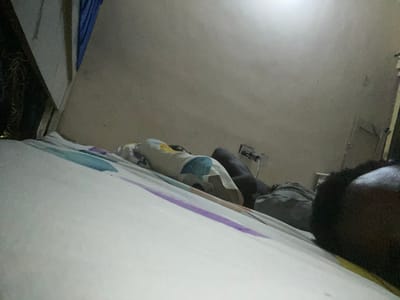 A person is lying on a bed with patterned sheets in a room. The person is lying on their side, facing away from the camera. They are wearing a light-colored shirt with a geometric pattern and dark-colored pants. The bed is covered with white sheets and a comforter with stripes of blue, purple, and orange. To the left of the bed, a blue curtain is visible. The wall behind the bed is a neutral color, and a light source is visible in the upper right corner, suggesting artificial lighting. The scene appears to be indoors, likely a bedroom, possibly at night given the lighting. The overall impression is a still, private moment.