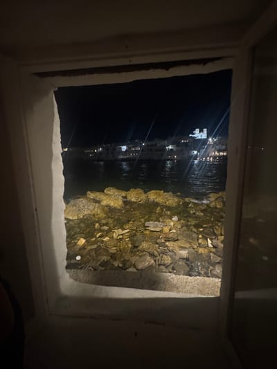 This image captures a nighttime view through an aperture, likely a window or opening, revealing a coastal scene in Naousa, Greece. The foreground is dominated by the rough, white interior of the aperture. Beyond this frame, a rocky shoreline is visible, with the sea gently lapping at the stones. In the distance, across the dark water, the lights of the town of Naousa are illuminated. A prominent white building, possibly a church with two bell towers, stands out against the backdrop of other buildings and lights. The water reflects the distant lights, creating streaks of light across the surface. The overall ambiance is one of quiet observation of the seaside town at night.