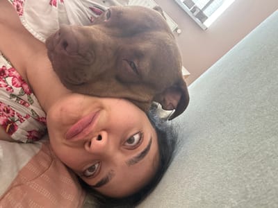This is a close-up, overhead selfie featuring a young woman and a dog. The woman is lying down, with her face close to the camera. She has dark hair and her eyes are wide open, looking upwards and to the left, with a surprised or playful expression. Her lips are pursed, as if she is about to kiss or make a funny face. She is wearing a white top with a floral pattern of pink and red flowers.
The dog, a brown pit bull or similar breed, is nuzzling against the woman's face and neck, partially obscuring her. The dog's eyes are partially closed, suggesting it is relaxed or sleepy. Its fur is short and appears to be a reddish-brown color.
The setting appears to be indoors, possibly a bedroom. The background shows a textured grey surface, likely a ceiling or wall, and a pink and white patterned pillow. A bit of a window with blinds is visible in the lower right corner, suggesting it might be daytime. The overall mood of the image is intimate and affectionate, capturing a moment between a woman and her pet. There is no visible text in the image.