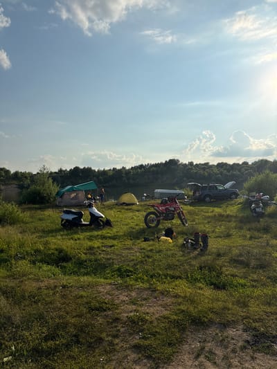 The image depicts a campsite on a grassy hillside overlooking a body of water, likely a river or lake, bordered by a dense line of trees. The sun is high in the sky, indicating it is daytime, and the weather appears to be clear with scattered clouds. The scene is set in Lan'shino, Russia.
Several objects are scattered across the landscape, suggesting an outdoor recreational gathering. There are at least two tents set up, one green and brown and another yellow. Multiple vehicles are present, including a dark-colored SUV with its doors open, a white van, and several motorcycles. One motorcycle, red and white, is prominently displayed in the foreground, leaning on its kickstand. Other objects include boots, bags, and what appears to be camping gear.
A few individuals are visible within the scene. One person is standing near the water with fishing gear. Another person is seated near a tent, and a few more are scattered around the campsite. The overall atmosphere suggests a leisurely outdoor activity, possibly camping and motorcycling. The bright sunlight and green foliage contribute to a feeling of a warm, pleasant day. There is no visible text in the image.