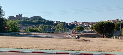 The image captures a view of the city of Oropesa, Spain, on a clear, sunny day. In the foreground, a dry, grassy field is visible, bordered by a road with red and white markings. A winding road curves through the middle ground, with a guardrail alongside it. Several traffic signs are present, including a red circular sign with a horizontal bar and a triangular yellow sign with two black triangles indicating a hazard. A person, appearing to be wearing dark clothing and a green item on their head, is seen bent over in the field. In the background, the city of Oropesa is situated on a hill. Prominent structures include a large castle with multiple towers and other buildings with terracotta roofs, characteristic of Spanish architecture. Lush green trees dot the landscape between the foreground and the city. Power lines are visible stretching across the upper portion of the image against the clear blue sky.
