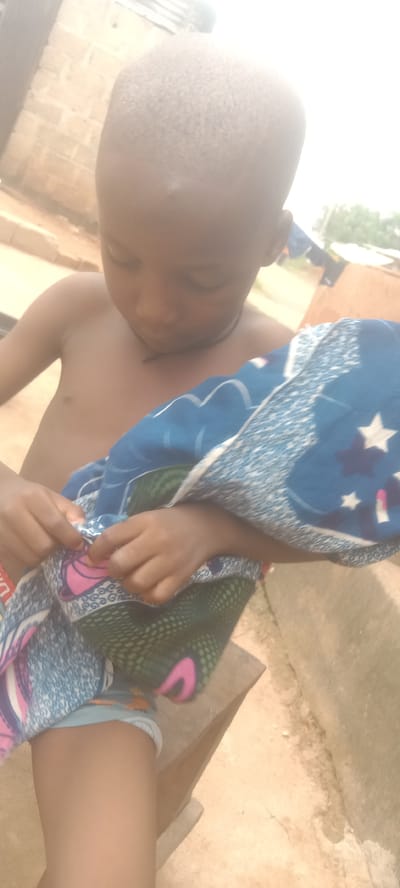 A shirtless young boy stands outside in Bende, Nigeria. He is holding a patterned blanket, looking down at it and focusing on something with his hands. The background includes a building and some laundry hanging to dry. The environment is bright and appears to be outdoors.