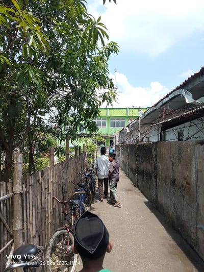 A narrow alleyway is framed by a weathered fence on one side and a concrete wall topped with barbed wire on the other. Bicycles lean against the fence. Two boys walk ahead, a man following behind them. A green building with windows is visible in the distance. The sky is partly cloudy. A third boy's head and shoulders are visible from behind in the foreground.