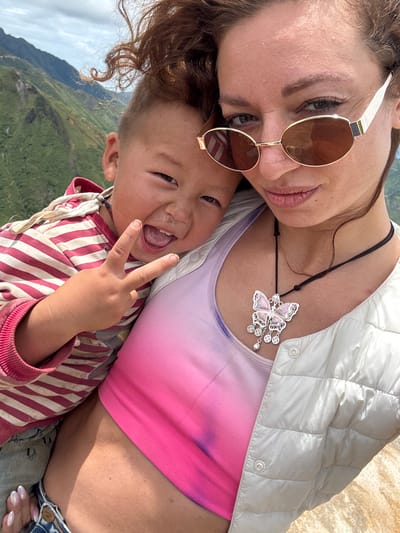 A woman and a child are in the foreground, with mountains in the background. The woman is wearing sunglasses, a pink and white tie-dye tank top, and a light gray puffer jacket. She is also wearing a necklace with a butterfly pendant. The child is wearing a red and white striped shirt and blue jeans. The child is making a peace sign with their fingers and smiling. The image appears to have been taken outdoors, possibly in a mountainous region. The mountains in the background are green and lush, suggesting a rural or natural setting. The sky is overcast with clouds. The location is identified as Bắc Yên, Vietnam.
