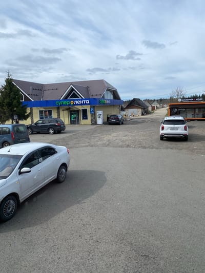 The image captures an outdoor scene at a commercial location in Seltso, Russia, under an overcast sky. In the foreground, an asphalt road occupies the right side, with a white car partially visible. To the left, a dirt parking area leads to two main buildings.
The most prominent structure is a supermarket with a blue facade and a distinctive brown, multi-gabled roof, featuring the large sign "СУПЕР O ЛЕНТА" (Super O Lenta). Parked in front of this building are a dark-colored SUV and a blue car. Further in the background, another, smaller building with an orange-brown facade houses a cafe. A white Hyundai Creta SUV, bearing the Russian license plate M539EE 174 (indicating Chelyabinsk Oblast), is parked near this cafe.
The activity depicted is typical for a commercial area, with cars parked for patrons visiting the stores or cafe. The cloudy sky suggests an overcast day, and some bare trees in the background imply it might be late autumn, winter, or early spring. A coniferous tree stands near the supermarket entrance.
Visible text includes: "СУПЕР O ЛЕНТА", a "P" parking sign, "OPEN", and "9-22" (indicating store hours) on the supermarket building. The cafe building displays "ШАВЕРМА & КОФЕ" (Shawarma & Coffee).
