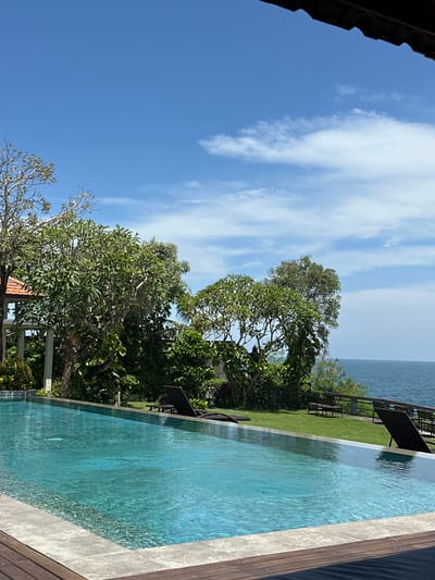 The image depicts a serene outdoor scene, likely a resort area, located in Uluwatu, Indonesia. In the foreground, a dark brown roof overhang partially frames the top right of the composition. Below this, the edge of a rectangular infinity pool with turquoise water is visible, featuring a light grey stone coping and a dark wooden deck surround on the left and bottom.
Beyond the pool, a well-maintained green lawn extends towards the horizon. Two dark-colored sun loungers are positioned on this lawn; one is located centrally behind the pool, and another is partially visible on the far right. A dense line of tropical trees and shrubs with vibrant green foliage forms a natural barrier behind the lawn. To the far left, a structure with terracotta-tiled roofs and light-colored columns is partially obscured by trees.
The vegetation gives way to a clear view of the open ocean, characterized by calm blue waters extending to the horizon. The sky above is bright blue with scattered white, wispy clouds, indicating clear weather. No individuals are present in the scene, and no overt activities are depicted. The overall impression is one of tranquility and natural beauty.