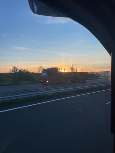 A red Scania semi-truck with a flatbed trailer, loaded with timber, is captured traveling on a multi-lane highway during sunset near Portel-des-Corbières, France. The truck moves from left to right, its headlights illuminated. The sun, a bright orange-yellow sphere, is visible at the horizon directly behind the truck, casting an orange hue across the lower sky that fades into light blue overhead. The highway features distinct white lane markings and a metal guardrail separating the lanes. Sparse trees and vegetation are visible along the distant horizon. The image is framed by a dark, irregular object in the upper and right sections, indicative of a vehicle's interior window frame from which the photograph was taken.