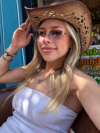 A young female subject is depicted from the chest up, seated outdoors in Tlaquepaque, Mexico. She has long, blonde hair with highlights and is wearing a light-colored, likely white, strapless top. She wears a woven straw cowboy hat with an openwork pattern and rimless sunglasses with purple-tinted lenses and gold-toned accents. Her left hand is raised, with fingers near the brim of her hat, revealing multiple rings on her left ring and middle fingers, and dark nail polish. On her left wrist, she wears two distinct bracelets: one composed of dark, spherical beads and another featuring flat, rectangular segments. A silver-colored necklace with an intertwined ring pendant (one silver, one gold) is visible around her neck. Her expression is a slight smile.
The background on the immediate left consists of a vibrant blue wall with a vertical white LED light strip. To the right and behind the subject, elements of a commercial beverage station are visible. This includes various bottles, a clear glass container, and a partially obscured sign with white text on a green and orange background, displaying "mojitos" and "margaritas." Above this area, decorative string lights with yellow, spherical elements (possibly lemons) are discernible. Strong sunlight illuminates the scene, casting distinct shadows on the subject's right arm and chest from an unseen overhead source, consistent with an outdoor or semi-outdoor restaurant or bar patio setting within Tlaquepaque. A dark brown surface, possibly a chair or table, is visible in the lower left quadrant.