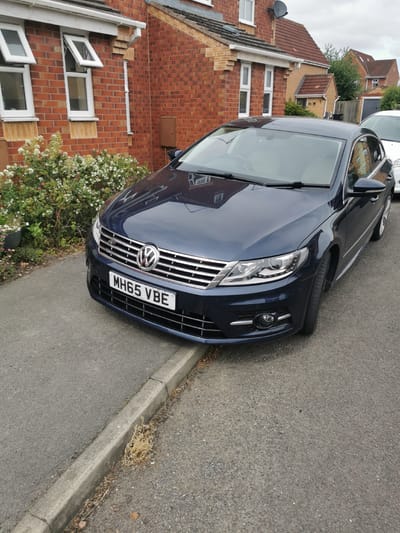 This is a medium shot, eye-level photograph of a dark blue Volkswagen CC parked on a residential street in Ripon, United Kingdom. The car, with its license plate "MH65 VBE" clearly visible, is positioned on the right side of the frame, partially obscuring a concrete curb and asphalt road. The front of the car, including its distinctive grille with the Volkswagen logo, headlights, and bumper, is in sharp focus.
To the left of the car, a paved walkway runs alongside a red brick house. The house features several white-framed windows, some of which are open. A small bush with white flowers and some pink blossoms is situated between the walkway and the house. In the background, other houses with red tiled roofs are visible, along with trees and a cloudy sky, suggesting an overcast day. The scene depicts a typical suburban residential area. No people are present in the image. The overall impression is of a quiet, ordinary street.