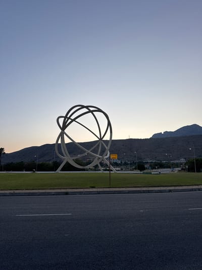 A wide-angle, eye-level shot captures a monumental spherical sculpture of intersecting white rings standing on a manicured grassy roundabout in Benidorm, Spain. The sculpture, appearing abstract and modern, is positioned centrally on the roundabout. Behind it, a barren, rocky mountain range stretches across the horizon under a clear, pale blue dusk sky. The foreground features a dark asphalt road with a white dividing line. In the middle ground, the roundabout is bordered by a curb and sidewalk, with streetlights and some trees visible. A yellow directional sign with Spanish text is mounted on a pole near the sculpture. The overall scene is serene and evokes a sense of an urban or suburban landscape at twilight.