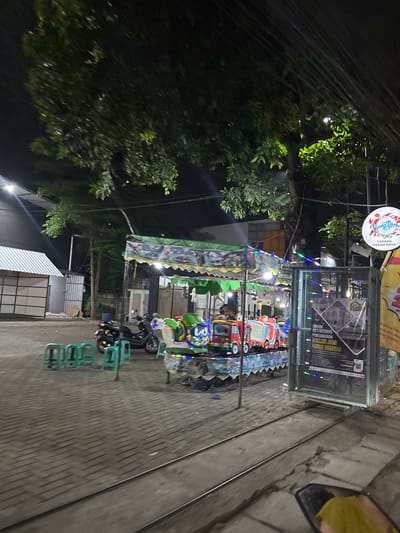 This image captures an outdoor scene at night, likely in South Tangerang, Indonesia. The primary focus is a small amusement ride area, possibly for children, with brightly colored toy vehicles. String lights are draped around the attraction, adding to the festive nighttime atmosphere. In the background, there are trees and some architectural elements, including a sign that appears to be for "Masjid Jami Nurul Hidayah" and mentions "Cabang Graha Raya," indicating a mosque branch. The ground is paved with patterned bricks, and there are metal tracks running along it, suggesting the amusement ride might follow these tracks. A black scooter is parked nearby, and several green plastic chairs are stacked, ready for visitors. The overall mood is casual and inviting, set against the backdrop of a clear night.