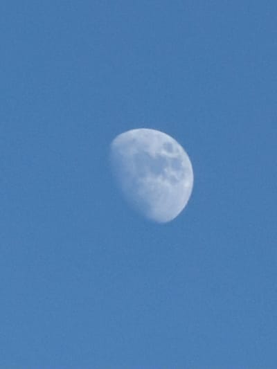 The image shows the moon against a clear blue sky. The moon is partially illuminated, with the visible portion appearing to be a waxing gibbous phase. The surface of the moon shows some craters and maria, which are darker, smoother areas. The sky is a uniform shade of blue, suggesting it is daytime. There are no buildings, landmarks, or other objects visible in the image, making it impossible to determine its specific location in Riverhead, United States of America.