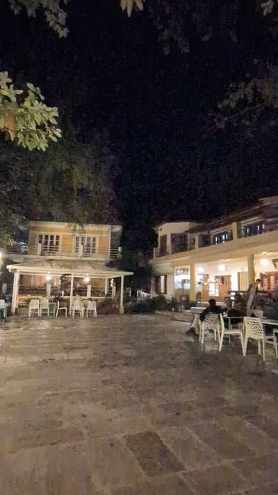 This nighttime image depicts an outdoor dining or lounge area, likely associated with guesthouses or cafes in Dharamkot, India. The scene features a paved ground on the right, bordered by dark, lush foliage on the left and in the foreground.

Two multi-story buildings are visible, illuminated from within. The larger building in the mid-ground is brightly lit, showcasing its interior through numerous windows and suggesting an active establishment. Closer to the foreground, a smaller building also features indoor lighting and a distinct outdoor seating area with a covered patio structure.

Numerous white or light-colored tables and chairs are scattered across the paved ground, some clustered around the buildings. A few individuals are discernible; one person is seated at a table in the outdoor section of the larger building, and other indistinct figures appear in the vicinity of the tables of the smaller building.

The time of day is clearly night, as indicated by the darkness of the surroundings and the artificial lighting emanating from the buildings and seating areas, which creates a warm and inviting glow. The activity suggests patrons are dining, socializing, or relaxing outdoors. The ambiance appears calm and peaceful. No visible text is legible.