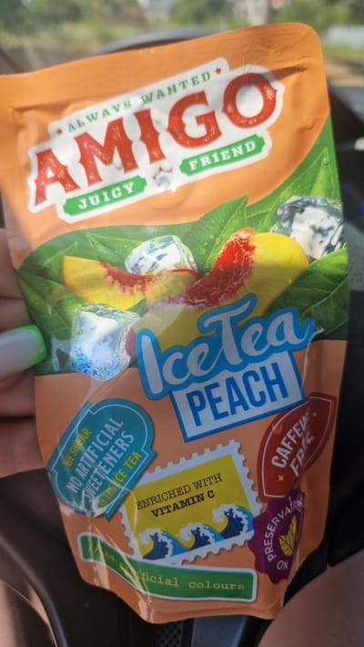 This is a close-up, high-angle shot of an orange pouch of "AMIGO" Peach Ice Tea, held by a person's hand. The pouch is predominantly orange with vibrant graphics of peaches, ice cubes, and green leaves, suggesting a refreshing beverage. Text on the pouch includes "AMIGO," "JUICY FRIEND," "ALWAYS WANTED," "Ice Tea PEACH," "CAFFEI FREE," "PRESERVAT ON," "ENRICHED WITH VITAMIN C," and "NO ARTIFICIAL SWEETENERS IN THIS ICE TEA," "nosaxificial colours." The lighting suggests it is daytime, possibly sunny due to the highlights and shadows. The background is blurred but appears to be an indoor setting with a dark surface, possibly a car interior or furniture. A person's thumb with a green-tipped fingernail is visible at the bottom right, holding the pouch. The image's focus is on the product packaging and its branding.
