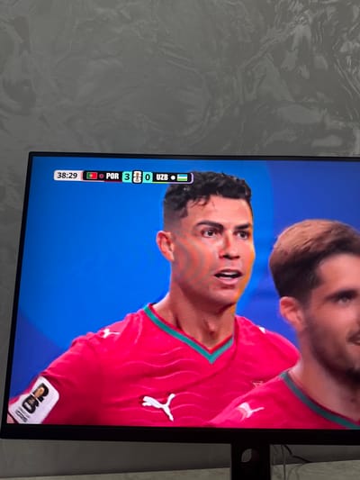 The image displays a close-up of a television screen showing a football match. On the screen, a scoreboard indicates the time is 38:29 and the score is POR 3 - 0 UZB. Two male footballers are visible. The player in the foreground, on the left, is wearing a red jersey with teal accents and appears to be Cristiano Ronaldo, based on his facial features. He is looking off to the side with an open mouth. To his right, another male footballer in a similar red jersey is partially visible. The background is a solid blue color. The television screen is placed on a stand or surface, and behind it, a wall with a textured, greyish finish is visible. The bottom of the television frame is also visible. There is no indication or visual information in the image that places the scene in Osh City, Kyrgyzstan.