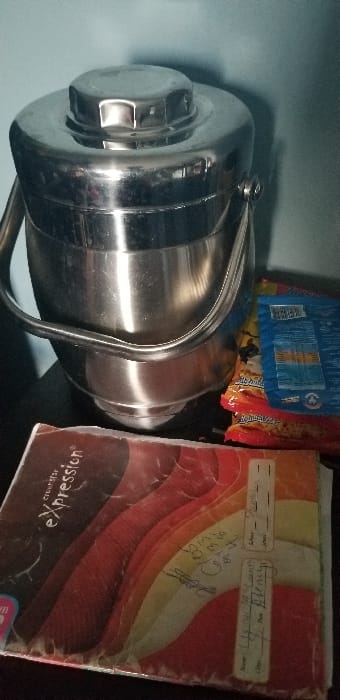 This image depicts a static indoor scene in a location identified as Ado-Ekiti, Nigeria. The primary subject is a large, cylindrical, stainless steel insulated food container or flask, positioned centrally. It features a reflective exterior, a prominent screw-on hexagonal lid, and a folding metal handle attached to its upper sides.

To the left and front of the container, a colorful notebook or exercise book is visible, resting flat. Its cover displays a "Creative eXpression" title and a design of wavy, layered bands in shades of dark brown, red, orange, yellow, and beige. Handwritten information, including labels for "Name," "Class," "School," and "Date," is present on the right side of the cover, with some illegible entries.

Behind the stainless steel container, several packaged items are stacked. At least two red-colored packages, possibly containing snacks, are discernible, with one partially revealing what appears to be "Beloxxi" branding. A blue-colored sachet or package is partially visible behind the red items.

All objects rest on a dark, flat surface, likely a table or shelf. The background consists of a plain, light blue-green wall. No individuals are present in the image, and no actions or interactions are depicted.