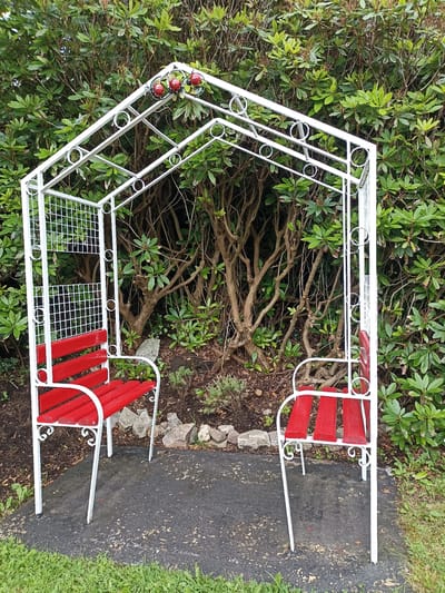 This is a full-shot, eye-level, outdoor photograph of a white metal garden arbor with two red benches. The arbor is positioned in front of a dense green shrubbery, likely a rhododendron bush, with visible branches and leaves. The ground beneath the arbor is a dark asphalt-like surface, with some gravel and small rocks bordering the shrubbery. To the left and right of the arbor, there is green grass.
The arbor has a decorative, arched structure with circular motifs and a diamond-shaped lattice on the left side. The two benches are made of white metal frames with red wooden slats for the seats. The bench on the left is positioned further back and partially obscured by the arbor's structure. The bench on the right is more visible. At the apex of the arbor, there are decorative red ladybugs with green leaf accents.
The scene appears to be set during the daytime, with diffused light suggesting an overcast or cloudy sky. There are no people visible in the image. The overall impression is that of a peaceful garden setting, perhaps a quiet spot for reflection or conversation in Annagry, Ireland. There is no visible text in the image.