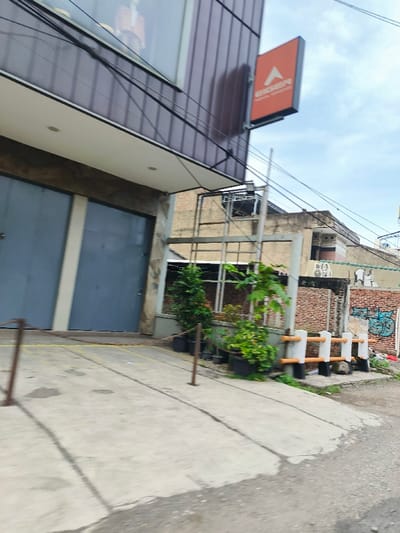 The image shows a street scene in Kecamatan Margahayu, Indonesia, a suburban area in the city of Bandung, located at the coordinates -6.964 latitude and 107.577 longitude. The image was taken from a street level perspective looking towards the front of a building. The building has two large garage doors painted grey, a brown awning and a large window. There is an orange sign with the word “AEROSPACE” in white lettering attached to the front corner of the building. The building is partially obscured by a metal fence, beyond which are plants and a brick wall with graffiti on it. The wall has several potted plants lined along the side of the building. The road surface is cracked and dirty, with yellow lines painted down the side.  The image is taken on a cloudy day, and the sky is grey. There are no people visible in the image.