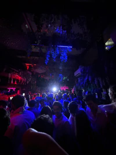 The image captures a densely crowded indoor space resembling a nightclub or bar, characterized by low-level, multi-colored lighting during evening hours. The environment is predominantly dark, illuminated by ambient blue and red light sources. Overhead and along walls, artificial green foliage, some adorned with blue string lights, forms part of the decor. A row of blue LED strip lights is visible on the upper ceiling. Red lighting fixtures are present on the left side of the frame. A bright white spotlight emanates from the background, center. An air conditioning unit is mounted on the upper right wall. In the background, a pink neon sign is partially legible, displaying "Welcome to Th...".
Numerous individuals, appearing to be young adults, are tightly packed throughout the foreground and midground. They are standing close together, engaging in social interactions or dancing. Their attire largely consists of light-colored shirts that reflect the colored ambient lighting. On the far left, one person is seen with an arm around another. Most faces are obscured by shadows, backlighting, or other individuals.
The specific location within Washington, United States, cannot be identified from the visual evidence in the image.