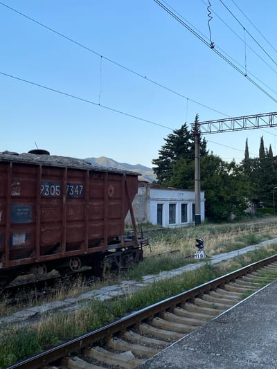 This image captures a train track area with a freight car in the foreground. The train car is rusty and appears to be an older model, with the numbers "2305 7347" visible on its side. It is parked on a track lined with concrete sleepers and gravel. Beyond the tracks, there is a low, white building with multiple windows and what appears to be a signal light labeled "M19" positioned near the tracks. Several tall, green trees stand behind the building, and in the distance, mountains are faintly visible. Overhead, power lines and a metal support structure for them dominate the upper left portion of the frame, against a clear blue sky. The lighting suggests it is daytime, likely late afternoon or early morning, given the soft light. The scene appears to be a quiet, perhaps disused, section of a railway line in Mtskheta, Georgia, with no people visible. The overall impression is one of stillness and a sense of a bygone era due to the aged appearance of the train car and the infrastructure.