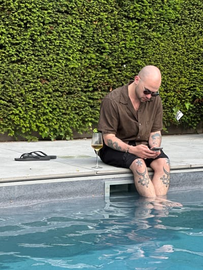 A bald, light-skinned man is seated next to a swimming pool with his legs submerged in the water. He is wearing sunglasses and a short-sleeved brown shirt. His arms and legs are covered in tattoos. He is holding a metallic object in his hands. A glass of white wine is on the ground next to him, along with a pair of black flip-flops. A thick green hedge forms a backdrop. The location is Cambridge, United Kingdom.