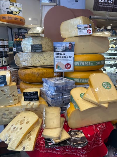 This image displays a diverse and abundant cheese counter in a shop, likely a specialty dairy store or deli, located in Noordwijk, Netherlands. The main subjects are various types of cheese, presented for sale on shelves and display surfaces.
A wide array of cheeses is visible, predominantly large blocks and wedges, most individually wrapped in clear plastic. These range in color from pale cream to deep yellow and orange, with some varieties featuring herbs or a marbled appearance. Prominently displayed are multiple blocks of "Graskaas Subliem B.O.B. 48+", advertised with a smiling man's picture on promotional signs. These signs, reading "SMAAK VAN HET VOORJAAR? PROEF ONZE GRASKAAS!" (Taste of Spring? Try our Grass Cheese!), show a price of €8.49 for 500 grams, mentioning its origin from Lutjewinkel, Noord-Holland.
Other identifiable cheeses include "Goudse Olijf-Tomaat" (€0.50), "Full Moon Kaas" from Denmark ("Thise fuldmåneost"), priced at €4.95 per 100 grams, and "Trommer Kaas," a type of holed cheese ("gatenkaas") aged 8 to 10 weeks, selling for €9.95. Some cheeses, like the "Full Moon Kaas," sit on a decorative red bandana-patterned cloth.
Beyond the cheeses, the display includes stacks of blue and white patterned paper napkins. In the background, other products are visible, such as a "Boska" brand fondue set box ("Super Cheesy") priced at €69.95, and jars of what appear to be spreads or dips. Signs for "KERSTPAKKETTEN & RELATIEGESCHENKEN" (Christmas Packages & Business Gifts) and "Zuivelhoeve Proef onze nieuwe dip!" (Taste our new dip!) suggest a broader range of products and seasonal offerings.
The scene depicts a well-lit, organized retail environment, highlighting the quality and variety of dairy products available. The activity is the presentation of cheese for sale, inviting customers to sample and purchase.