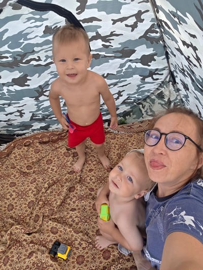 An overhead, medium shot captures three subjects inside a tent with a camouflage pattern on the interior walls. On the left, a young boy with blond hair and wearing red shorts smiles at the camera. He is holding a blue and pink toy. In the foreground, to the right, a woman with glasses and red hair is taking a selfie. She is sticking her tongue out and has a baby with blue eyes in her arms. The baby is holding a green toy car. A patterned blanket covers the floor of the tent, and on it, two toy cars, one black and one yellow, are visible. The image is taken inside a tent, likely for recreational purposes.