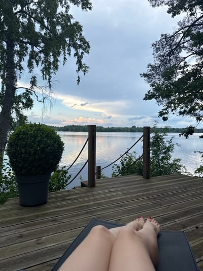 The image depicts a serene lakeside scene, likely in Priedesmuiža, Latvia, during what appears to be late afternoon or early evening. In the foreground, a person's legs are visible, resting on a dark-colored lounge chair on a wooden deck. Their toenails are painted red. The deck itself is constructed from weathered wooden planks.

To the left of the deck, a large potted green plant, possibly a boxwood, stands in a dark planter. Beyond the deck, a calm lake stretches out, reflecting the sky. The opposite shore is lined with a dense forest of green trees. The sky above is a mix of blues and grays, with scattered clouds, some illuminated by what seems to be the setting sun. Two wooden posts with ropes attached are present on the deck, serving as a railing. Lush green foliage is visible on the right side of the deck, partially obscuring the view of the lake. The overall atmosphere is one of peaceful relaxation by the water.