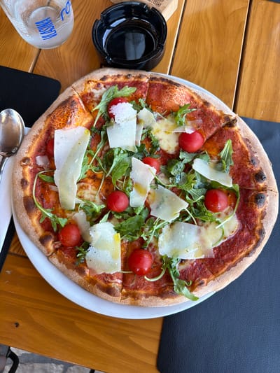 A high-angle, close-up shot captures a Margherita pizza on a white plate, positioned on a wooden table at a restaurant in Ulcinj, Montenegro. The pizza is topped with tomato sauce, melted cheese, cherry tomatoes, arugula, and shaved parmesan. A glass bottle of Vlasina water, a black ashtray, and a silver spoon are also visible on the table. The scene is lit by natural light, casting a warm glow on the food and surroundings. The background is blurred, focusing attention on the pizza. The overall impression is one of a delicious and appealing meal enjoyed in a relaxed setting.