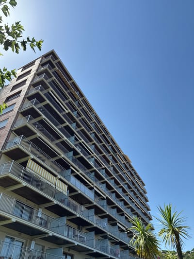 The image shows a modern, multi-story building with a distinctive facade of repeating balconies and angled windows. The building is viewed from a low angle, looking upwards towards a clear blue sky. Green foliage from trees is visible in the foreground and to the right of the building, partially obscuring the upper floors. The building's exterior appears to be constructed from brick on the lower visible sections, transitioning to a darker material and glass with metal railings on the upper levels. Some balconies have retractable awnings with yellow and white stripes.
The scene suggests a bright, sunny day, likely in a warm climate, given the presence of palm-like fronds. There are no people visible, and the building appears residential or possibly commercial. The architectural style is contemporary, with a strong emphasis on repeating geometric patterns. The perspective and clear sky evoke a sense of openness and urban living. The location cues of Sant Feliu de Guíxols, Spain, reinforce the impression of a Mediterranean setting. There is no visible text in the image.