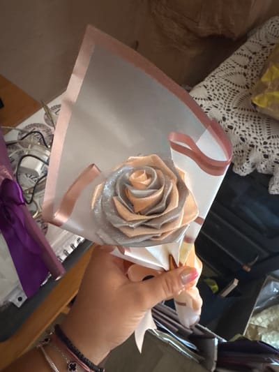 A person's hand is holding a bouquet wrapped in transparent paper, adorned with pink ribbons. The bouquet contains a single, intricately folded rose made of satin fabric, with silver glitter accents. In the background, a white crocheted doily is visible on a surface. To the left, a purple metallic ribbon or fabric is partially visible. The environment suggests an indoor setting, possibly a home or a craft space, with various items in the background including what appears to be storage containers and other crafting materials. The lighting suggests it is daytime. The image does not provide any specific visual information about the city of Sredets, Bulgaria.