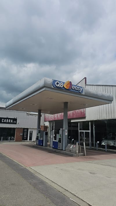The image shows a Q8 easy gas station on a cloudy day. The gas station is located in Boussu-lez-Walcourt, Belgium. The main structure of the gas station consists of a large canopy supported by pillars. Underneath the canopy are several fuel pumps, some of which are blue and grey. To the left of the gas station, there is a building with a sign that reads "CABRA.cc". Further to the left, there is a storefront with a window displaying what appears to be bicycles. To the right of the gas station, through a large glass window, a white car is visible, with the word "OCASION" on its rear. The sky is filled with dark, heavy clouds. The ground in front of the gas station is paved with asphalt and a reddish-pink surface.