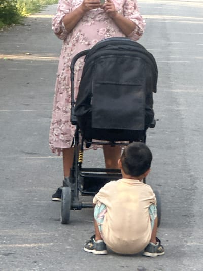 In this image, a young boy in a light tan t-shirt and patterned shorts is crouched next to a stroller. He appears to be interacting with the stroller, possibly playing with it or inspecting it. The stroller is black with a metallic frame. To the left of the stroller, a person wearing a light pink floral dress is visible from the waist down, with their arm extended towards the stroller. The scene appears to be outdoors on a paved surface, likely a street or sidewalk, given the texture of the ground. The lighting suggests it might be daytime, possibly late afternoon or early evening, with soft shadows. The overall mood is casual and domestic, capturing a moment of everyday life. No visible text is present in the image. The context of Тайгараев, Kyrgyzstan, places this scene in a specific geographic and cultural setting.