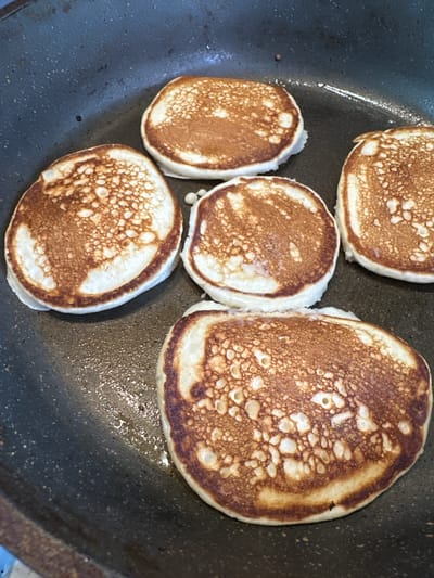 Five golden-brown pancakes are cooking in a black non-stick frying pan. The pancakes have a porous texture and are unevenly browned, with some lighter patches. The pan contains a small amount of shimmering cooking oil. The background is the dark interior of the frying pan. The scene is consistent with food being prepared in a kitchen environment, possibly in a home or restaurant in Yaroslavka, Russia. There are no people or other objects visible in the image.