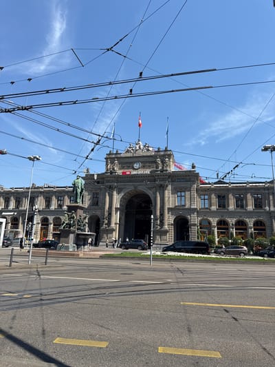 The image depicts the exterior of Zurich Hauptbahnhof (Zurich Main Station), a prominent railway station located in Zurich, Switzerland. The station's grand architecture, characterized by classical columns and an arched entrance, is visible under a clear blue sky with scattered clouds. Overhead, a complex network of overhead power lines for trams or trains crisscrosses the frame, obscuring parts of the sky and the upper facade of the building.

In the foreground, a wide asphalt road features tram tracks and yellow road markings. Several vehicles, including cars and a black van, are present on the road and the sidewalk. A statue of a man in historical attire stands in front of a fountain to the left of the station's main entrance. Several individuals are seen walking on the sidewalk and near the fountain, some appearing to be tourists or commuters. The station's facade displays signage, including the SBB CFF FFS logo and a clock, indicating its function as a railway hub. The overall scene suggests a bustling urban environment on a bright, sunny day.