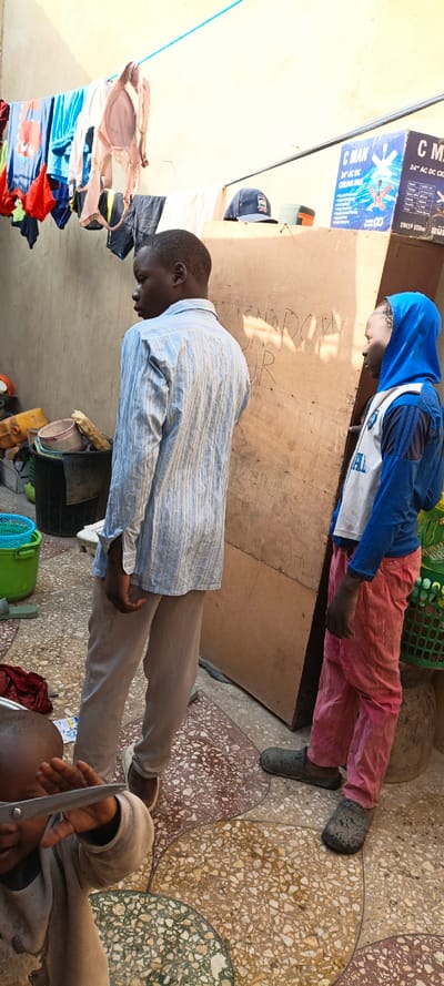 The image portrays an outdoor domestic scene in Kaura Goje, Nigeria. Three individuals are present on a multi-colored terrazzo-style tiled floor. In the foreground, a child, partially cropped, holds a pair of silver-handled scissors. Standing center-left, a male in a light blue and white striped long-sleeve shirt and light brown trousers faces right, away from the camera. To his right, a younger male, seen in profile, wears a blue hooded long-sleeve shirt with white and blue chest detailing, pink trousers, and dark slip-on shoes, also facing right.
Behind the two standing males is a large, vertical light-brown wooden panel bearing dark, handwritten graffiti, including "SNAROWY JR". To the left, various containers are stacked, including a black bucket, a white bucket, and yellow jerrycans. Above, a blue rope clothesline extends across the upper frame, draped with assorted garments, including t-shirts and a light-colored bra. In the upper right, a "C MAN 24" AC/DC CEILING FAN" cardboard box is visible on a surface, next to a dark blue baseball cap. The background features light-colored walls, and the scene is illuminated by bright daylight.