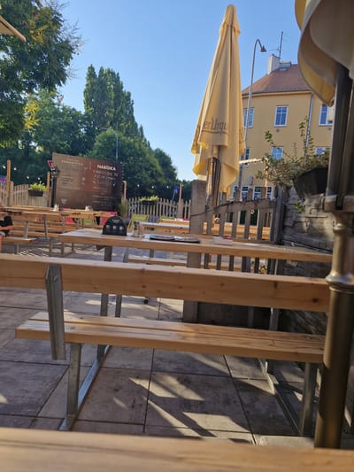 The image depicts an outdoor seating area with wooden picnic tables and benches, likely at a restaurant or cafe. A large, folded yellow umbrella is visible, branded with "er Urqu" which suggests a connection to the Pilsner Urquell brewery. In the background, a light yellow building with white-framed windows stands under a clear blue sky, indicating a sunny day, likely in the afternoon given the shadows.

A prominent sign, written in what appears to be Czech, lists "NABÍDKA" (Offer) with various drinks and food items. Some of the text is legible, mentioning "NOVÉ ŽIVÉ A ZELENÉ LIMONÁDY" (new live and green lemonades), "BOMBASTICA", and "MATCHA MALINA" (matcha raspberry). The scene is set in Brno, Czechia, and the overall atmosphere suggests a relaxed, casual dining or drinking environment.

The perspective is from a seated position, looking across the tables and towards the background building and trees. The sun casts distinct shadows from the wooden structure and possibly from surrounding trees onto the paved ground. There are no people clearly visible in the foreground, but the setup implies a place ready for patrons. The presence of a street sign in the background further grounds the location as an urban or semi-urban setting.