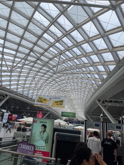 The image shows the interior of a modern building with a distinctive geodesic dome-like roof structure made of white beams and translucent panels, allowing natural light to filter through. The perspective is looking upwards and diagonally, emphasizing the architectural grandeur of the ceiling.
In the foreground and mid-ground, several people are visible. Some are standing and appear to be in a queue or waiting area, possibly for security checks or at a service counter, given the presence of security scanners. Others are walking. The overall atmosphere suggests a public space like an airport, train station, or large exhibition hall.
Various signs and banners are displayed throughout the scene, some featuring advertisements and others providing information. One prominent sign shows an image of a man and text that appears to be in Chinese, possibly promoting a brand or service. Another banner has the text "美团闪购 马上到" which translates to "Meituan Flash Sale Arrives Immediately." Other text visible includes "SPOKESMAN" on a white t-shirt, and "安全检查" meaning "Security Check."
The lighting suggests it is daytime, with the bright sky visible through the roof. The setting appears to be indoors, likely a busy public transit hub or commercial center in Panyu District, China, given the Chinese text on the signs. The people are dressed casually, and their expressions are not clearly discernible due to the image resolution and perspective. The image captures a dynamic moment in a well-lit, architecturally interesting public space.