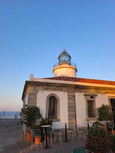 The image shows the Faro de Sant Sebastià lighthouse, a prominent landmark located in Palafrugell, Spain. The lighthouse is a white, cylindrical structure topped with a glass lantern room. It is situated on a white building with a red-tiled roof. The building has stone accents and arched windows with metal grates. In front of the building, there are several outdoor tables and chairs, suggesting a cafe or restaurant setting. Two potted olive trees are visible, and a person is walking away from the camera towards the sea in the background. The sky is a clear blue, indicating a sunny day. The lighting suggests it is either early morning or late afternoon, as the side of the lighthouse is illuminated with a warm glow. The text "FARO DE S. SEBASTIAN" is inscribed on the building.