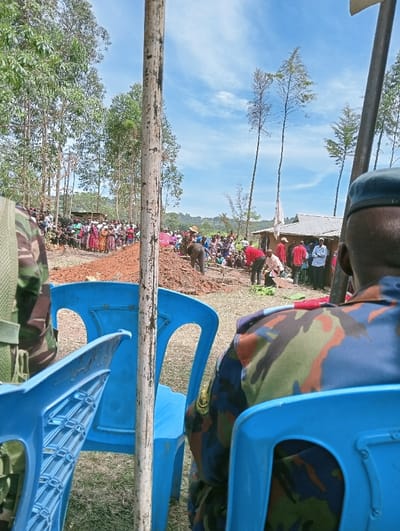 The image depicts an outdoor community gathering in a rural setting under a bright, sunny sky in Kipkarren, Kenya. In the immediate foreground, two individuals wearing military-style camouflage uniforms are seated on blue plastic chairs, with their backs to the viewer, observing the scene. A prominent, weathered wooden pole stands vertically in the center foreground, partially obscuring the view of the gathering.

In the mid-ground, a large crowd of people is assembled on reddish-brown, unpaved ground. Many attendees are standing, observing an area where several individuals are actively engaged in digging or moving earth, suggesting communal labor, a groundbreaking ceremony, or potentially a funeral. Piles of freshly disturbed soil are visible in this central area. The crowd comprises people of various ages, many wearing brightly colored clothing.

The background features a verdant landscape with numerous tall, slender trees, some resembling eucalyptus, under a clear blue sky dotted with scattered white clouds. Simple structures, possibly houses or shelters with corrugated iron roofs, are visible nestled amongst the trees in the distance. The bright sunlight indicates it is daytime, likely late morning or early afternoon. The scene conveys an atmosphere of collective activity and community engagement. There is no visible text in the image.