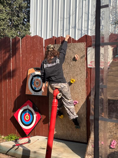 An individual, viewed from the back, is engaged in rock climbing on a vertical wooden panel (approximately 2.5 feet wide) affixed to a reddish-brown picket fence in an outdoor recreational area within the Menashe Regional Council, Israel. The climber wears a camouflage bandana, a black long-sleeved shirt with "payback is death" in white script across the shoulders, and camouflage pants, with black socks or footwear visible on their left foot. Their right arm is extended, gripping the top edge of the wooden panel, while their left foot is positioned on a yellow climbing hold. Other colored climbing holds (one orange, two yellow, two pink) are attached to the wooden panel.
In the foreground, a tall, red cylindrical standpipe or fire hydrant-like structure with a top valve partially obscures the climber's lower right side. To the left of the climber, two archery targets are mounted on the reddish-brown fence: an upper rectangular target and a lower diamond-shaped target, both featuring concentric rings and a "10" bullseye, with "GEOLOGIC" branding. A small red and black bow with an arrow rests on the concrete ground beneath the lower target. The background includes a white corrugated metal building to the right, obscured in part by a reflective surface, and green foliage from trees to the left. Sunlight indicates a daytime setting, casting shadows on the concrete surface. A sign with Hebrew text ("בדו") is visible on the fence to the right of the climbing panel.